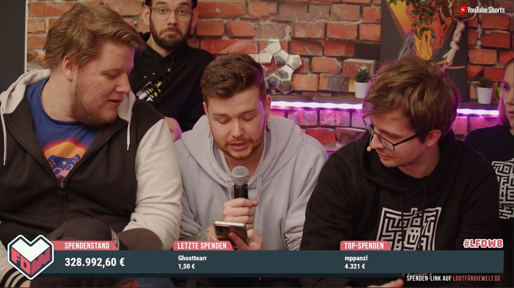 Wir sehen einen Screenshot von Loot für die Welt 8. Drei junge Männer, alle weiß mit braunen Haaren sitzen vor einer Backstein-Wand. Zwei tragen Mikros am Kopf, einer hält ein Handmikro. Er schaut auf sein Handy und liest etwas vom Handy vor. Die beiden anderen schauen rechts und links neben ihm auch auf den Screen. Unten sehen wir das Overlay im Design von Loot für die Welt mit dem Logo links. Daneben steht "Spendenstand: 328.992,60 €", "Letzte Spenden: Ghosttearr 1,50 €" und "Top-Spenden: mppanzl 4.321 €"