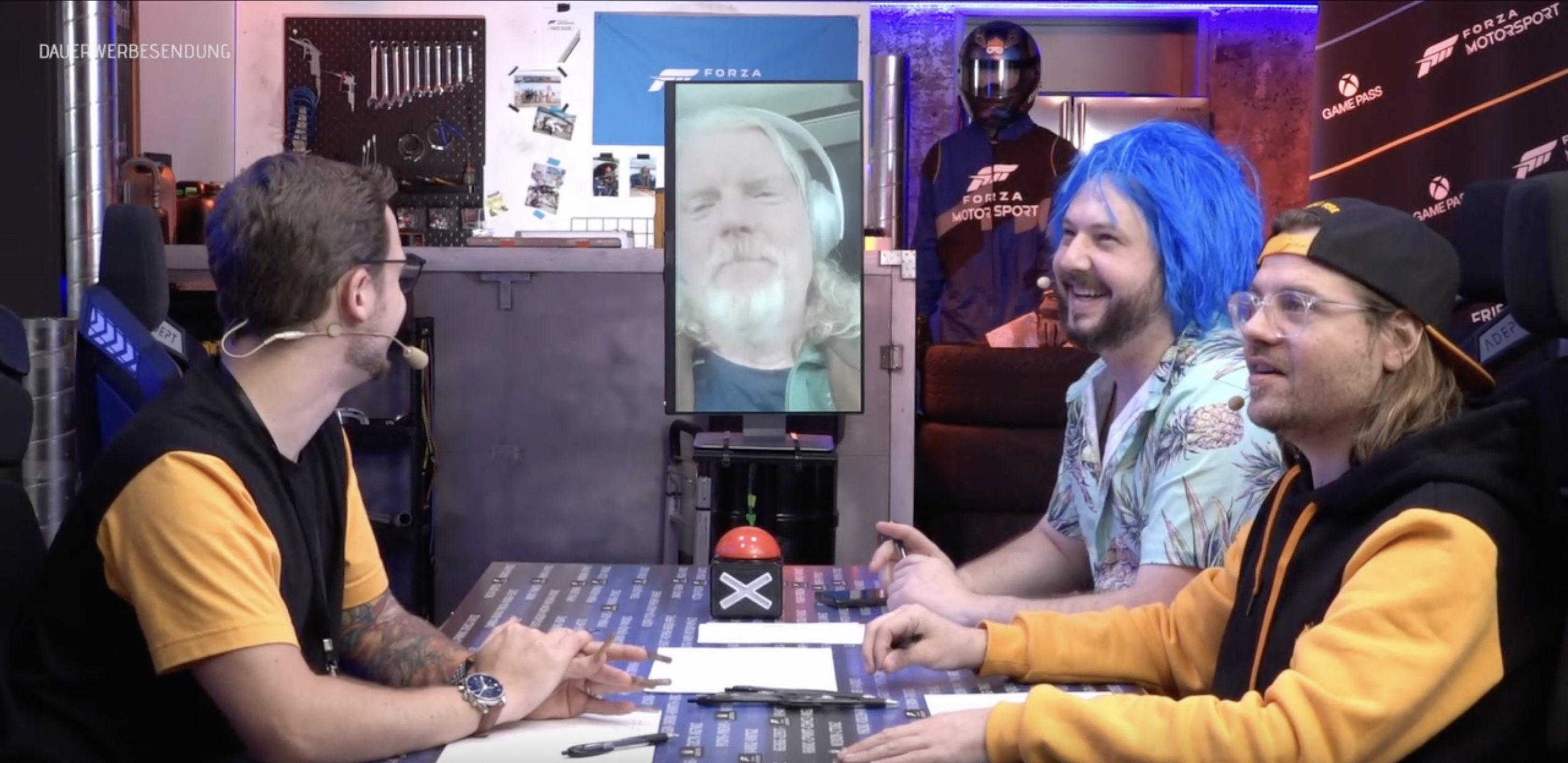 Sep, FisHC0p und Chris aus dem Friendly Fire Team sitzen an einem Tisch und vor ihnen liegen Stifte und Papier. Zentriert im Hintergrund ist über einen vertikalen Monitor Dan Clancy zu sehen, der über eine Videokonferenz dazugeschaltet wird