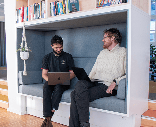Zwei Kollegen sitzen nebeneinander mit Laptops auf dem Schoß und unterhalten sich