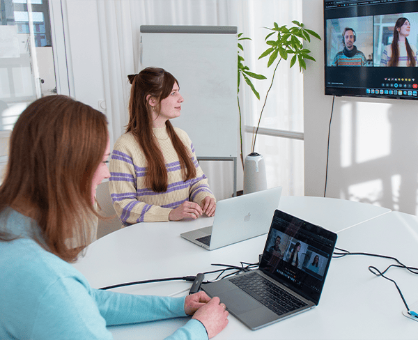 Zwei Kolleginnen sitzen in einem Meetingraum und haben eine Videokonferenz.